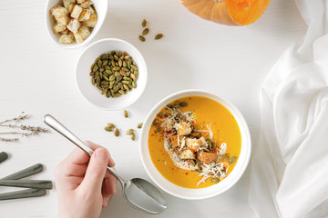 Pumpkin soup, a piece of pumpkin, seeds and crackers lie on a white table. Nearby is a white towel and serving utensils. Male hand holds a spoon.