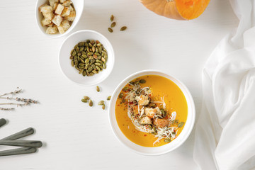 Pumpkin soup, a piece of pumpkin, seeds and crackers lie on a white table. Nearby is a white towel and serving utensils.

