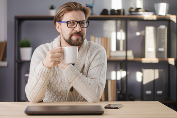 Happy bearded man in glasses and knitted sweater sitting at table with cup of coffee and looking at camera. Mature person taking break during work at home.