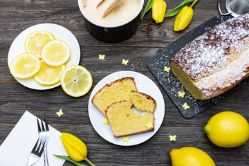 Teller mit zwei Scheiben Zitronenkuchen auf einem Holztisch von oben fotografiert, daneben Kuchen, Tulpen, Zitronen, Zitronenscheiben, Zucker und Dekoration