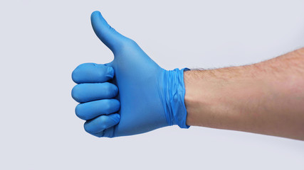 Male hand in a blue medical glove show a raised thumb up during quarantine. Isolated on a gray background in the studio. Coronavirus Concept. Stay at Home.