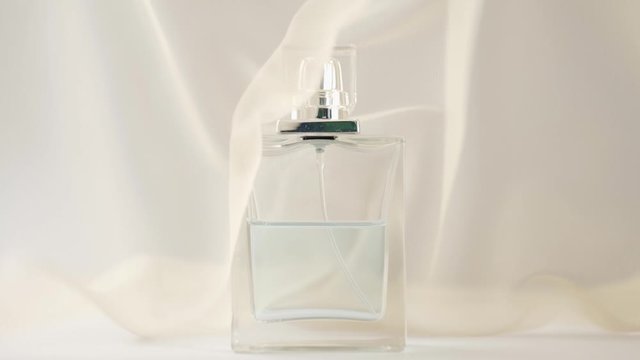 Flat surfaced bottle with cyan perfumes or essential oils is on the white table. Beige fabric flutters around and waves in the air around the bottle. Concept of aroma and smell. Close up. Slow motion