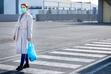 Donna con mascherina verde, cappotto chiaro , borsa nera  e una busta blu  cammina sulle strisce...