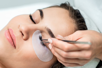 Cosmetologist hand doing eyelash extension procedure by tweezers on eyelashes of woman, close up. - obrazy, fototapety, plakaty