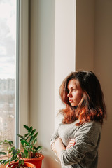 A brunette girl with long hair and a homemade gray sweater stands against an empty wall next to the window and touches a homemade green plant zamiokulkas