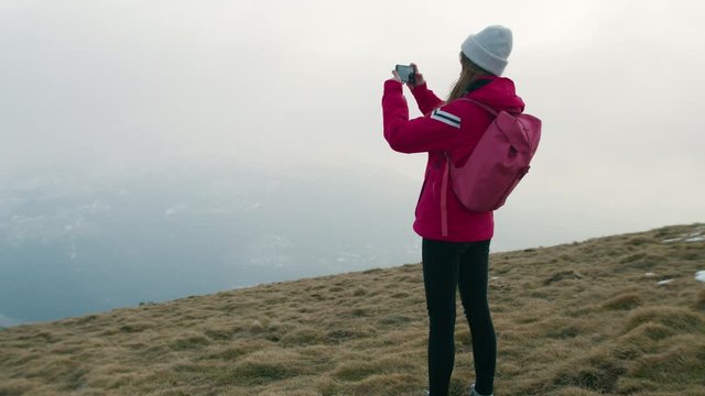 Back View of Young Female Hiker in Red Jacket Standing on Top of Mountain and Taking Video or Panoramic Shot on Smartphone. Free Girl Hiking in Nature and Exploring Natural World. Slow Motion Shot