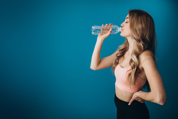 Side view of attractive young girl with curly blond hair drinking fresh water over blue studio background. Fitness woman in sport outfit refreshing after workout.