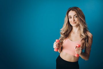 Beautiful sporty woman in activewear standing in studio with two dumbbells. Young girl with blond hair doing exercises for arms. Isolated over blue background.