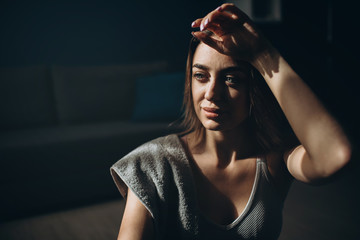 Portrait of attractive young lady sitting on floor with towel on shoulder and feeling tired after workout. Sporty woman taking break during exercises at home.