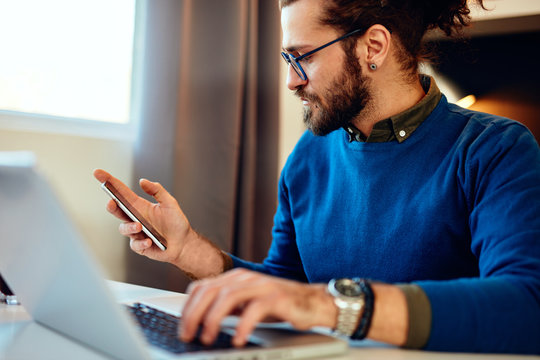 Attractive Caucasian Bearded Entrepreneur Sitting In His Home Office, Using Smart Phone And Taking A Break From Work. Work From Home Concept.