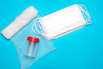 Close up of medical masks and a roll of white sterile gauze bandage with a can for passing a urine test with a red lid on a blue background