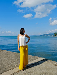 Positive woman walking at the harbor on vacation. Mountains on background. Cheerful, smiling, lucky lady in white blouse, yellow trousers and sunglasses, outdoors at sea port. Sunny summer day.