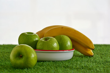 Bodegon de fruta sonbre un cesped de color verde y un fondo blanco. En la imagen hay fresas, arandanos, manzanas, platanos.