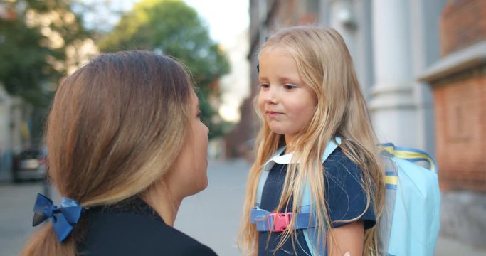 Close up view of little adorable child hugging her mother before leaving school at street. Young attractive mother talking and waving to her daughter that running to class in morning.