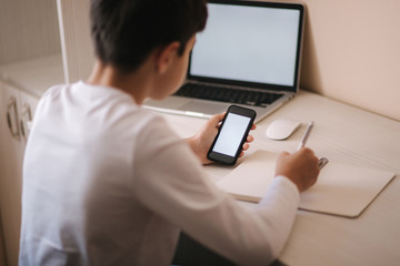 Schoolboy study at desk in his room. Boy use laptop and smartphone to writing in notebook. Books and tablet on the table. Study home during qurantine