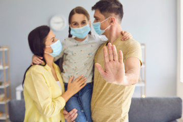 Family in medical masks on their faces looks at the camera while standing in a room at home. Father hand forward up concept of protecting a family of people from the threat of virus infection.