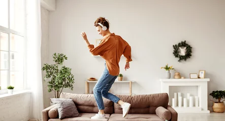 Gordijnen Vrolijke vrouw die naar muziek luistert en danst op een zachte bank thuis in een vrije dag. © JenkoAtaman