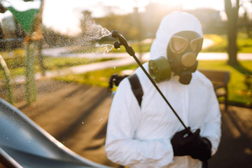 Man wearing protective suit disinfecting the playground in the sun with spray chemicals to preventing the spread of coronavirus, pandemic in quarantine city. Covid -19. Cleaning concept.