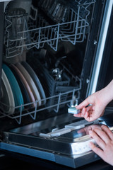 Hand of a young woman putting a tablet in a dishwasher detergent box close-up with utensils loaded
