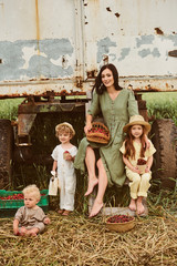 Beautiful young caucasian mother with her children in a linen dress with a basket of strawberries gathers a new crop and has fun with the children near the trailer