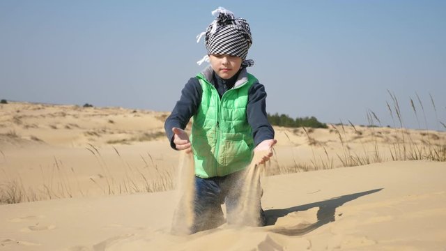 Cheery boy in a keffiyeh, jacket and jeans playing sith sand in a spring desert