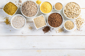 Selection of whole grains in white bowls - rice, oats, buckwheat, bulgur, porridge, barley, quinoa, amaranth, on white wood background