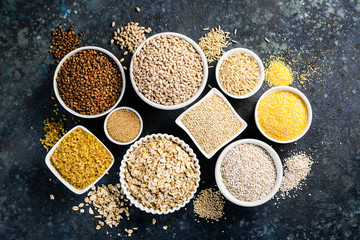 Selection of whole grains in white bowls - rice, oats, buckwheat, bulgur, porridge, barley, quinoa, amaranth on dark background