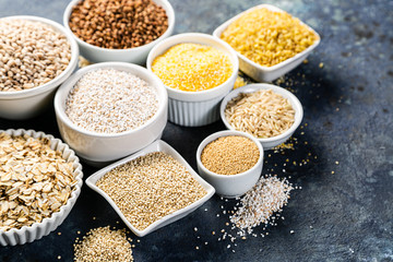 Selection of whole grains in white bowls - rice, oats, buckwheat, bulgur, porridge, barley, quinoa, amaranth on dark background