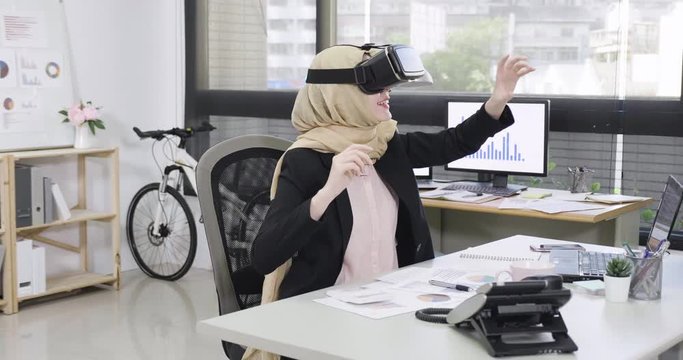 Using VR technologies. Confident young malay woman in virtual reality headset pointing hands catching in air while sitting at working place in office. smiling muslim lady worker having fun in break.