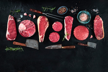 Various cuts of meat, shot from above on a black background with salt, pepper, rosemary and knives, with a place for text