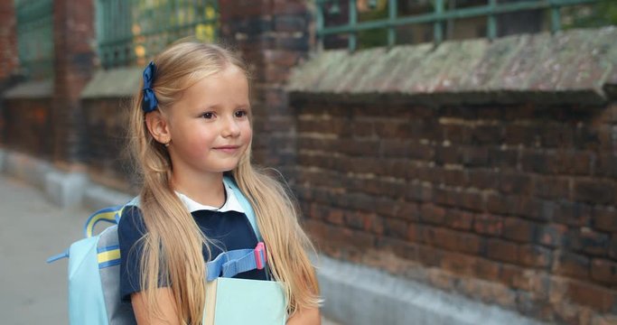 Little cute girl carrying book while walking at street. Pretty small child with long blond hair and bag on her back going slowly to school. Concept of children and education. Outdoors.