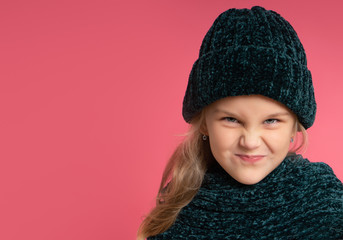 Little blonde kid dressed in green warm scarf and hat. She is smiling, looking naughty, posing against pink studio background. Childhood, fashion, advertising concept. Close up, copy space