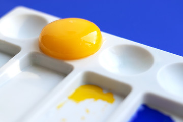 White palette with watercolor paints and brushes. Blue background. Instead of yellow, the egg yolk is used. Creative design and art. Top view and copy space