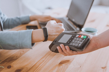 Woman working on laptop, using terminal for contactless payment via smartwatch for her order in cafe. New technology of NFC payment