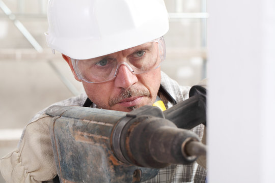 man using an electric pneumatic drill making a hole in wall, professional construction worker with safety hard hat,, gloves and protective glasses. inside construction site