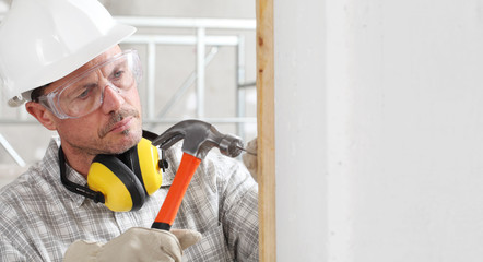 man construction worker with the hammer nails the plank to the wall wear gloves, hard hat, glasses and hearing protection headphones, at interior building site