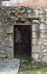 Old door with a rusty handle in a natural stone fence and a tiled roof. Treat sun falling through a big tree.