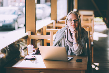Fototapeta premium A woman sits at a wooden table in a cafe and uses a smartphone. There is a laptop on the table. The girl looks through chat, blogs. The woman holds the phone and looks at the viewer
