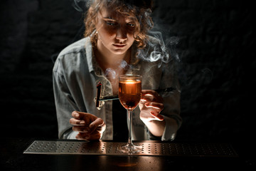 young woman at bar holds tweezers with smoking wand near glass of cocktail.