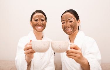 Two friends with mud masks on their faces drink tea. Spa relaxation, facial skin care, skin cleansing and treatment. Part of a series with mud mask