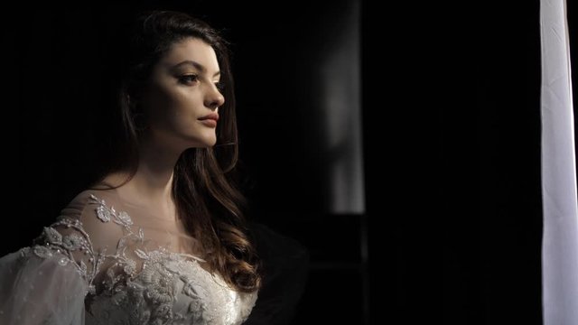 long haired brunette in white lacy wedding dress with transparent sleeves and neckline stands in darkness slow motion closeup