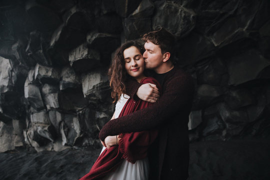 Wedding in Iceland. A young guy and a girl in a white dress and a red sweater are hugging near the black rock