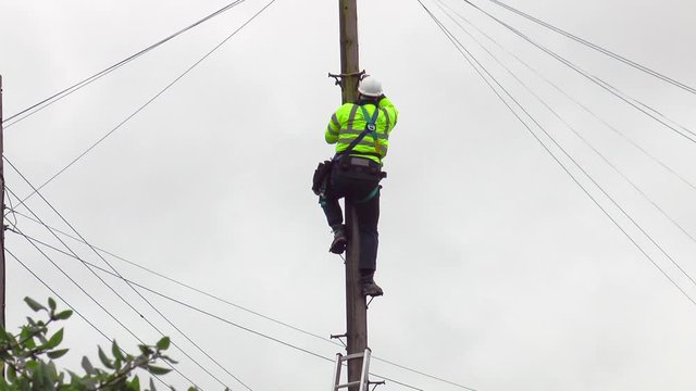 An unidentifiable telephone engineer wearing a safety helmet and high visibility jacket, loosening safety ropes and carefully climbing down a telegraph pole, with cables in all directions.