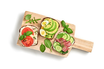 Open sandwiches with vegetables, avocado, tomato, mozzarella, boiled egg and soft cheese. Homemade sandwich with ham, cucumber, radish sprouts on wooden board isolated on white, top view