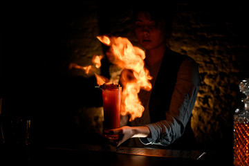 Woman bartender holding glass with cocktail sprinkles on it and masterfully make fire.
