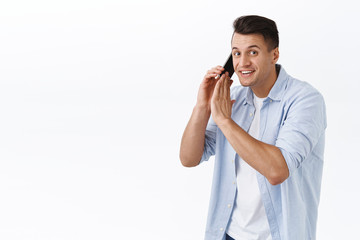 Sorry cant talk right now. Portrait of smiling handsome man busy talking on phone, hold mobile phone and cover dynamic to say something to you, having conversation, white background