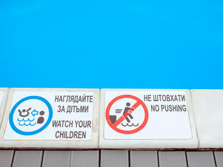 The edge of the swimming pool with the table with information "Watch you children" and "No pushing" in English and Ukrainian language. Sport, safety, recreation and relax concept.