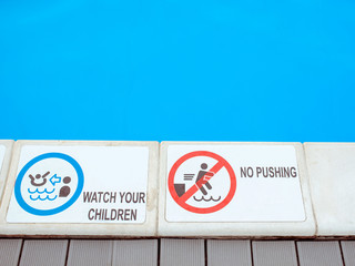 The edge of the swimming pool with the table with information "Watch you children" and "No pushing" in English language. Sport, safety, recreation and relax concept.