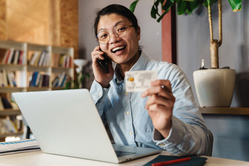 Image of joyful asian man talking on cellphone and holding credit card