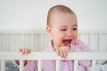 Baby in beautiful pink checked pajamas. The child woke up and cried a lot. A child is standing in its white crib.